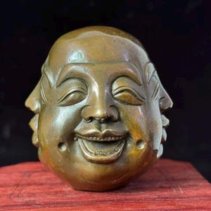 Könnte beinhalten: Eine bronzefarbene Skulptur eines mehrgesichtigen Buddha-Kopfes. Die Skulptur hat ein lächelndes Gesicht mit geschlossenen Augen und einem friedlichen Ausdruck. Die Skulptur steht auf einer roten Holzoberfläche vor dunklem Hintergrund.