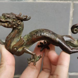 Könnte beinhalten: Bronze-Drachenfigur mit aufwendigen Details. Der Drache ist in einer dynamischen Pose dargestellt, mit gebogenem Körper und ausgestreckten Beinen. Die Bronze hat eine dunkle, gealterte Patina. Ein dekorativer Artikel.