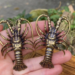 May include: Two bronze-colored lobster-shaped earrings. The earrings have detailed claws, legs, and antennae. The lobster bodies are textured, and the earrings are held in a hand against a blurred green background.