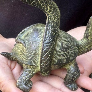May include: A bronze-colored turtle figurine with a snake wrapped around its body. The turtle has a detailed shell pattern and is held in open hands. The snake's body is textured, and the overall design is antique-inspired.