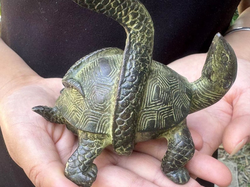 May include: A bronze-colored turtle figurine with a snake wrapped around its body. The turtle has a detailed shell pattern and is held in open hands. The snake's body is textured, and the overall design is antique-inspired.