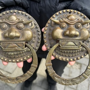 May include: Two antique bronze door knockers. Each knocker features a detailed, stylized face with an open mouth and a ring handle. The round backplates are adorned with a circular pattern of small studs.