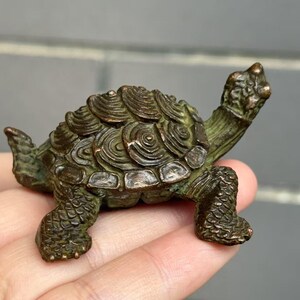 May include: A small, detailed bronze turtle figurine. The turtle has a textured shell with concentric circle patterns and detailed legs and head. The figurine is a dark bronze color with hints of green.