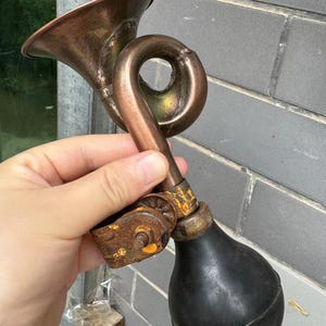 Puede incluir: Un claxon de bicicleta antiguo de lat&oacute;n con una pera de goma negra. El claxon tiene un dise&ntilde;o curvo y una campana acampanada. El metal muestra signos de edad y desgaste, con un soporte oxidado.