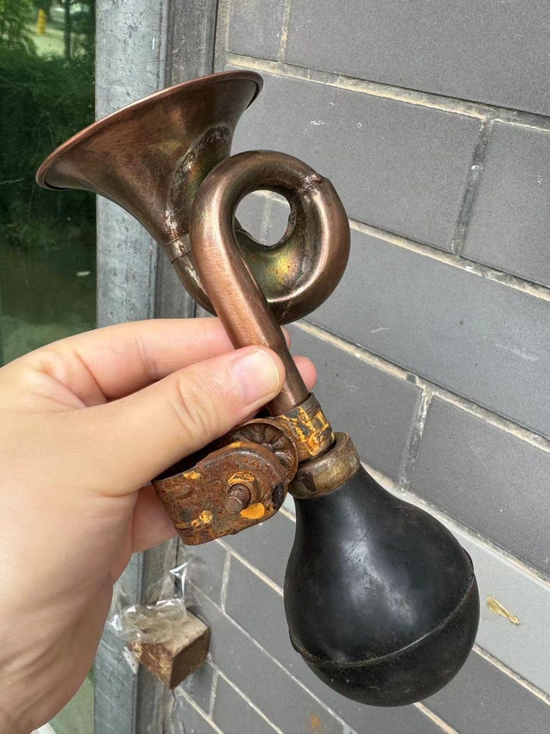 Puede incluir: Un claxon de bicicleta antiguo de lat&oacute;n con una pera de goma negra. El claxon tiene un dise&ntilde;o curvo y una campana acampanada. El metal muestra signos de edad y desgaste, con un soporte oxidado.