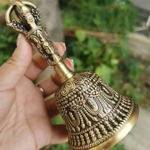 May include: Ornate brass bell with detailed carvings. The bell features a decorative handle with a figure and intricate patterns on the bell's body. The bell is held against a blurred green background.