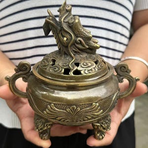 May include: A bronze-colored incense burner with a detailed dragon design on the lid. The burner has ornate handles and feet, and a decorative floral pattern around the base. The lid features cloud-shaped cutouts.