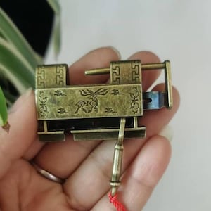 May include: An antique brass-colored padlock with intricate engraved designs. The lock features a rectangular body with decorative patterns and a long, slender key attached to a red string. The lock is held in a hand, suggesting its size and detail.