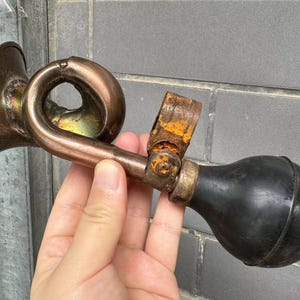 Puede incluir: Un claxon de bicicleta antiguo de color cobre con una pera de goma negra. El claxon tiene un dise&ntilde;o curvo y un soporte met&aacute;lico oxidado. El claxon se sostiene en una mano.