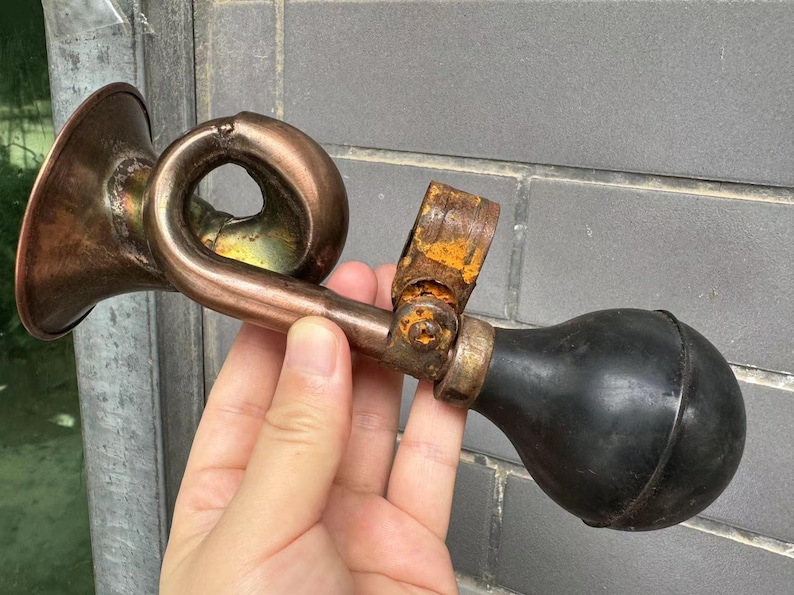 Puede incluir: Un claxon de bicicleta antiguo de color cobre con una pera de goma negra. El claxon tiene un dise&ntilde;o curvo y un soporte met&aacute;lico oxidado. El claxon se sostiene en una mano.
