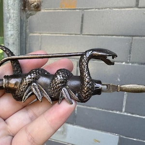 Puede incluir: Candado de color bronce con un diseño detallado de serpiente enrollada alrededor del cuerpo. La cabeza de la serpiente forma la parte superior del candado, con una llave insertada en el lateral. El candado tiene una estética vintage.