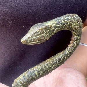 May include: A bronze-colored, snake-shaped decorative object. The snake's head is detailed with scales and a small eye. The body curves in an elegant arc. The object has a weathered, antique appearance, with hints of green and gold.