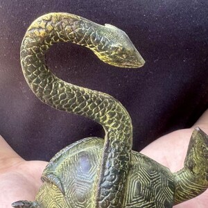 May include: A bronze sculpture of a turtle with a snake wrapped around its shell. The snake's head is raised, and the turtle's shell has a geometric pattern. The sculpture has a weathered, antique appearance.