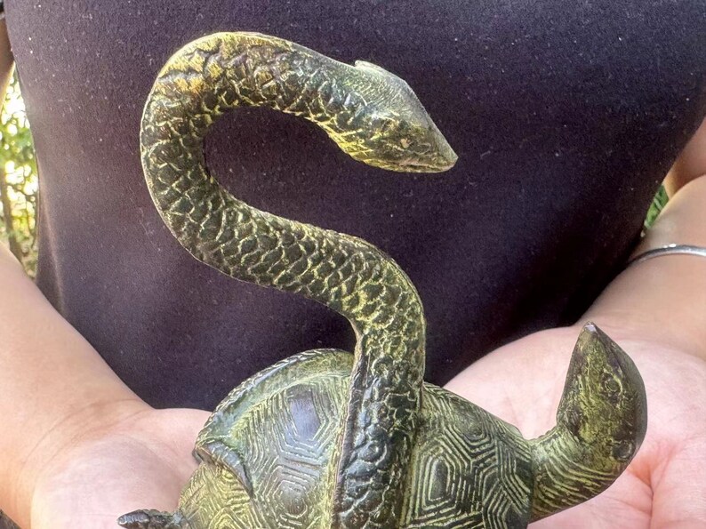 May include: A bronze sculpture of a turtle with a snake wrapped around its shell. The snake's head is raised, and the turtle's shell has a geometric pattern. The sculpture has a weathered, antique appearance.