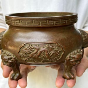 Puede incluir: Quemador de incienso de bronce con un relieve de dragón. El recipiente tiene un borde estampado y está sostenido por tres patas con cabeza de león. El cuerpo del quemador presenta un diseño de dragón detallado, lo que se suma a su apariencia ornamentada. Un artículo decorativo tradicional de inspiración asiática.