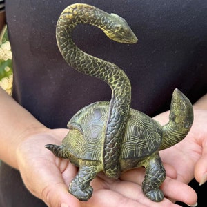 May include: A bronze-colored sculpture of a turtle with a snake wrapped around its body. The turtle has a detailed shell pattern and the snake's head is raised. The sculpture is held in a person's hands.