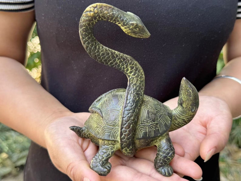 May include: A bronze-colored sculpture of a turtle with a snake wrapped around its body. The turtle has a detailed shell pattern and the snake's head is raised. The sculpture is held in a person's hands.