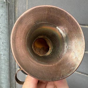 Puede incluir: Un embudo de metal color cobre con un asa peque&ntilde;a. El embudo tiene una abertura ancha y acampanada y un pico estrecho. El metal tiene una superficie texturizada con signos de desgaste. El embudo est&aacute; sostenido por una mano.