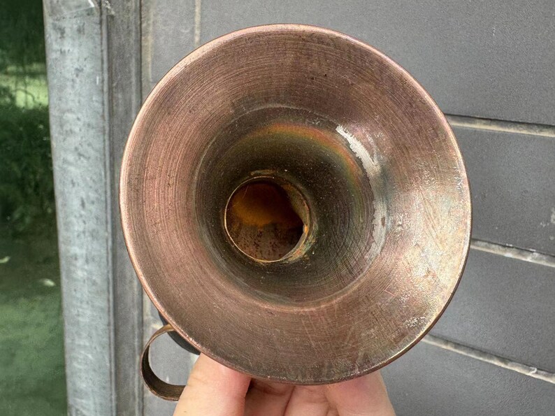 Puede incluir: Un embudo de metal color cobre con un asa peque&ntilde;a. El embudo tiene una abertura ancha y acampanada y un pico estrecho. El metal tiene una superficie texturizada con signos de desgaste. El embudo est&aacute; sostenido por una mano.