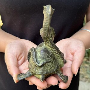 May include: A bronze turtle sculpture with a detailed shell pattern and an elongated neck. The sculpture is held in a person's hands, showcasing its intricate design and aged patina. The turtle's head is raised, adding to its unique aesthetic.