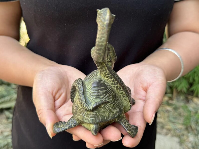 May include: A bronze turtle sculpture with a detailed shell pattern and an elongated neck. The sculpture is held in a person's hands, showcasing its intricate design and aged patina. The turtle's head is raised, adding to its unique aesthetic.
