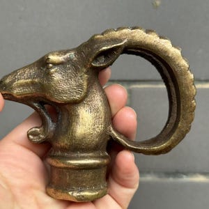 Op de afbeelding: Een bronzen flesstopper met een gedetailleerd ramshoofd ontwerp. De stopper heeft een getextureerd oppervlak en een gebogen handvat. De stopper wordt tegen een grijze achtergrond gehouden.