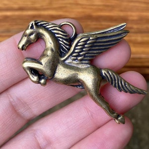 May include: A bronze-colored Pegasus pendant, a mythical winged horse, with detailed wings and a loop for hanging. The pendant is in a dynamic pose, suggesting flight, and is likely for jewelry or crafting.