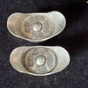 Puede incluir: Dos objetos plateados en forma de lingote con caracteres chinos. Cada uno tiene un diseño circular central con texto y un botón central en relieve. Los lingotes están sobre una superficie oscura y texturizada.