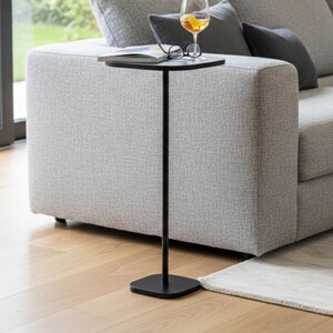 May include: A tall, slender black side table with a thin vertical post and a square base. The table features a flat, circular top surface that holds a glass of amber liquid with a citrus peel garnish, a pair of eyeglasses, and an open book.