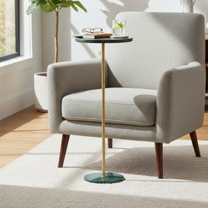May include: A light gray upholstered armchair with dark wood legs. A small side table with a green marble top and brass pole stands next to the chair. A glass of water and a book rest on the table.