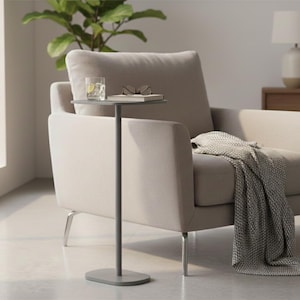 Pode incluir: Uma sala de estar moderna com uma poltrona bege e uma manta cinza. Uma pequena mesa lateral com um copo de água e óculos. Uma luminária de chão e uma planta também são visíveis.