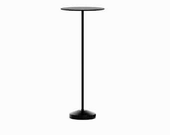 May include: A tall, slender black metal stand featuring a thin vertical pole, a flat circular top surface, and a weighted, conical base. The entire structure is finished in a solid matte black colour, providing a stable and minimalist display.