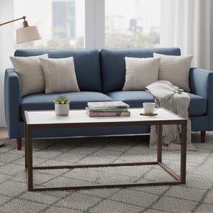 May include: A blue sofa with beige cushions and a white coffee table with a brown metal frame. A small potted plant, books, and a coffee cup are on the table. A floor lamp and a rug are also visible.