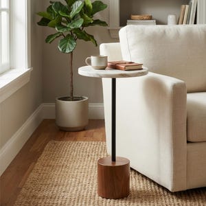 May include: A marble-topped side table with a black metal stem and a wooden base. A mug and a book rest on the table. A potted plant and a beige sofa are in the background. The table is on a woven rug.