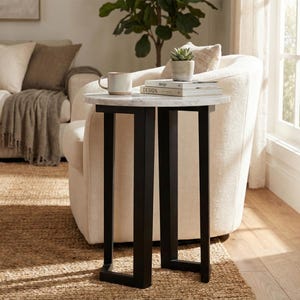 Pode incluir: Uma mesa lateral redonda com um tampo de mármore branco e uma base de metal preto. A mesa apresenta uma caneca, uma pequena planta suculenta num vaso e dois livros empilhados com a palavra "design" impressa nas capas, sobre a superfície.