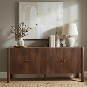 Könnte beinhalten: Ein Sideboard aus dunklem Holz mit vier Türen und Messingknöpfen. Auf der Oberseite stehen eine Lampe mit beigem Schirm und eine Vase mit getrockneten Zweigen. An der Wand hinter dem Sideboard hängt ein Gemälde in neutralen Farben.