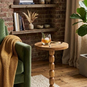 May include: A round wooden side table with a pedestal base composed of stacked, rounded wooden spheres. The table features a smooth, natural wood grain finish and supports a glass containing an amber liquid, a large ice sphere, and a sprig.