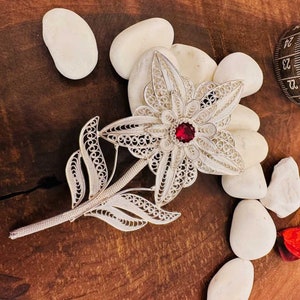 Puede incluir: Broche de flores de filigrana plateada con un centro de piedra roja. El broche presenta pétalos calados detallados y un tallo con hojas. El broche se exhibe sobre piedras blancas contra una superficie de madera. Mide aproximadamente 10 cm de largo.
