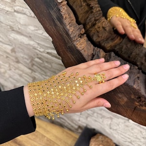 May include: A gold hand chain bracelet with a ring and dangling beads. The bracelet is made of a delicate, open-weave design with square and circular links. The ring features a floral design. The jewelry is displayed on a hand.
