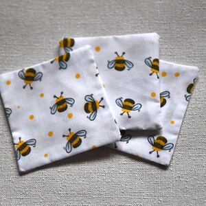 May include: Three white square fabric items featuring a repeating pattern of cartoon bees and yellow dots. The bees have black and yellow striped bodies, blue wings, and yellow antennae. The items are arranged on a light grey surface.