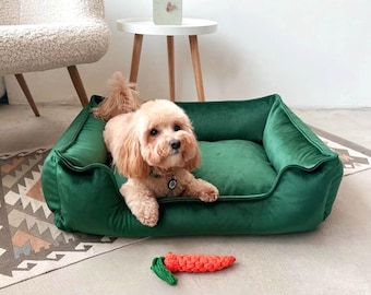 May include: A small, light brown dog lies on a plush, emerald green dog bed. A carrot-shaped dog toy is next to the bed. The bed is rectangular with raised sides. The dog wears a collar with a tag.