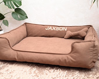 May include: A brown rectangular dog bed with a raised back and sides. The word "JAXSON" is embroidered on the back. A bone-shaped pillow rests inside the bed. The bed is made of a soft, corduroy-like material.