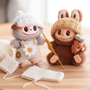 Puede incluir: Dos muñecos de peluche, uno gris y otro marrón, cada uno con un gorro de punto. El muñeco gris tiene un gorro blanco y un peto con una margarita. El muñeco marrón sostiene un ganchillo y una zanahoria. Piezas de ganchillo en primer plano.