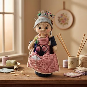 May include: A handmade crochet doll with a gray hat adorned with pins. The doll wears a pink apron with pockets holding scissors and other craft supplies. The doll is a whimsical organizer for sewing or knitting notions.