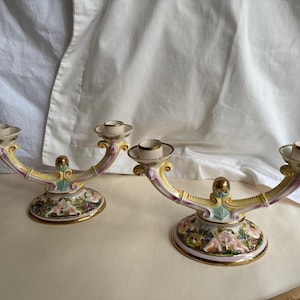 Peut inclure: Une paire de candélabres en porcelaine dotés de deux bougeoirs chacun, reliés par une structure incurvée jaune et rose avec des bordures dorées. Les bases circulaires sont ornées de motifs floraux colorés et de chérubins en relief.