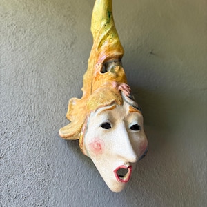 May include: A ceramic wall mask featuring a tall, pointed yellow hat with textured details. The face has a pale, crackled finish with pink-tinted cheeks, dark eye sockets, and an open, red-rimmed mouth. The sculpture has a pointed chin and nose.