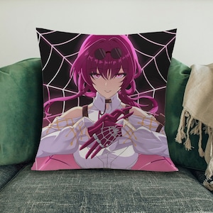 Könnte beinhalten: Ein rosa-weißes Kissen mit einem schwarzen Spinnennetz-Design. Eine Frau mit pinken Haaren und einem weißen Shirt befindet sich in der Mitte des Kissens. Sie trägt eine schwarze Halskette und eine schwarze Sonnenbrille. Sie hält ihre Hände vor sich, und ihre Finger sind zu Krallen gekrümmt. Die Frau hat einen ernsten Gesichtsausdruck.