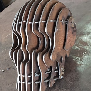 May include: A metal sculpture of a skull made from multiple layers of metal. The sculpture is rusted and has a rough texture. The skull is made up of several pieces that are connected together with rivets.