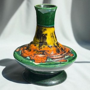 Puede incluir: Un jarrón de cerámica hecho a mano con un diseño único. El jarrón presenta una base y borde verde oscuro, una sección media amarilla y naranja con detalles en negro y un cuello acampanado. El jarrón mide aproximadamente 20 cm de alto.