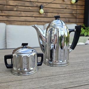 Può includere: Una caffettiera e una zuccheriera abbinata in cromo lucido con manici e pomelli del coperchio in plastica nera. Entrambi i pezzi presentano un design scanalato verticale sul corpo. La caffettiera ha un beccuccio curvo e un coperchio alto.